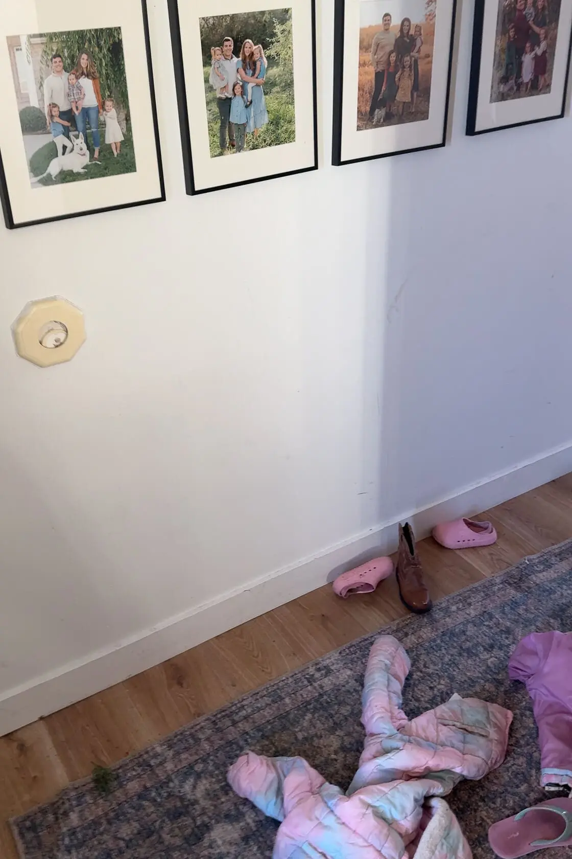 A hallway wall featuring family photo frames hung in a neat row above a cluttered floor with a pink jacket, mismatched shoes, and a rug. The plain white wall shows wear and tear, setting the stage for a home makeover.